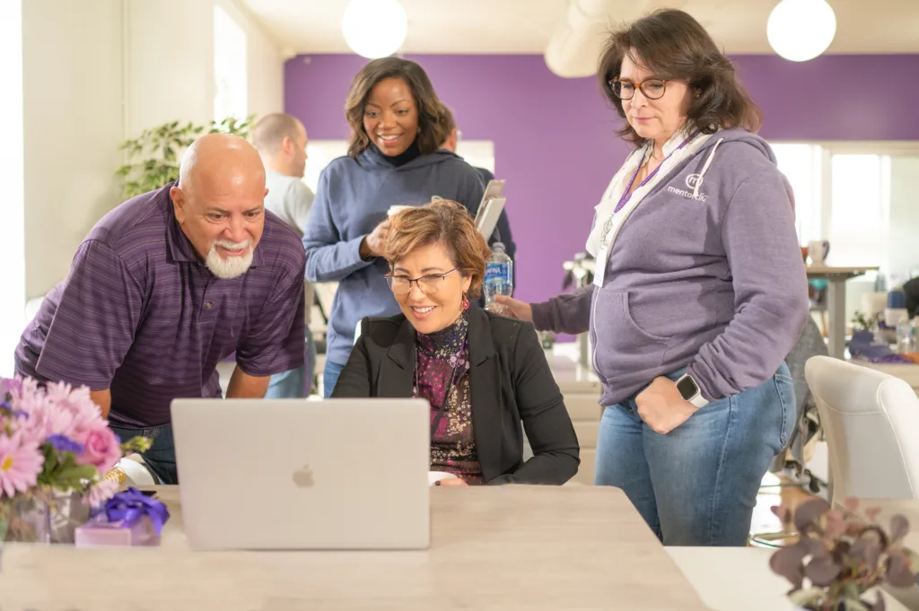 Four colleagues collaboratively looking at a laptop in a brightly lit office decorated with purple accents and fresh flowers on the table.