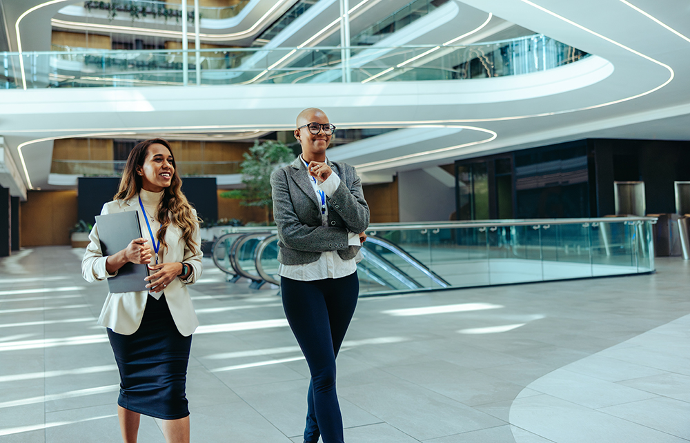 Two confident female financial professionals walk through a modern office building. They are dressed in business attire, capturing the essence of women thriving in a professional business setting.