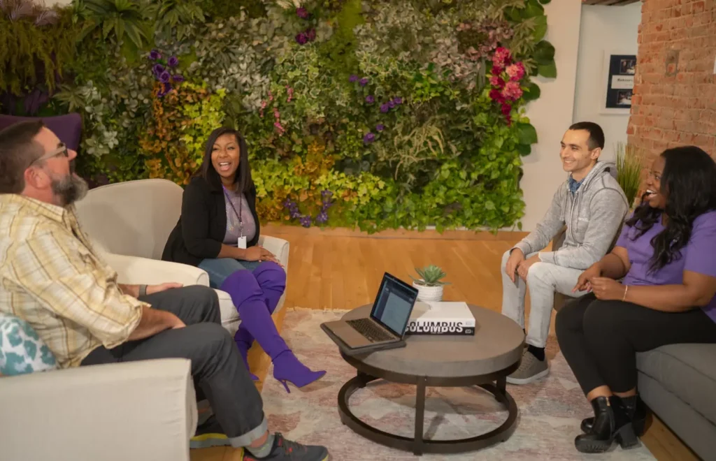 Coworkers having a casual group discussion in a modern office lounge with a green plant wall.