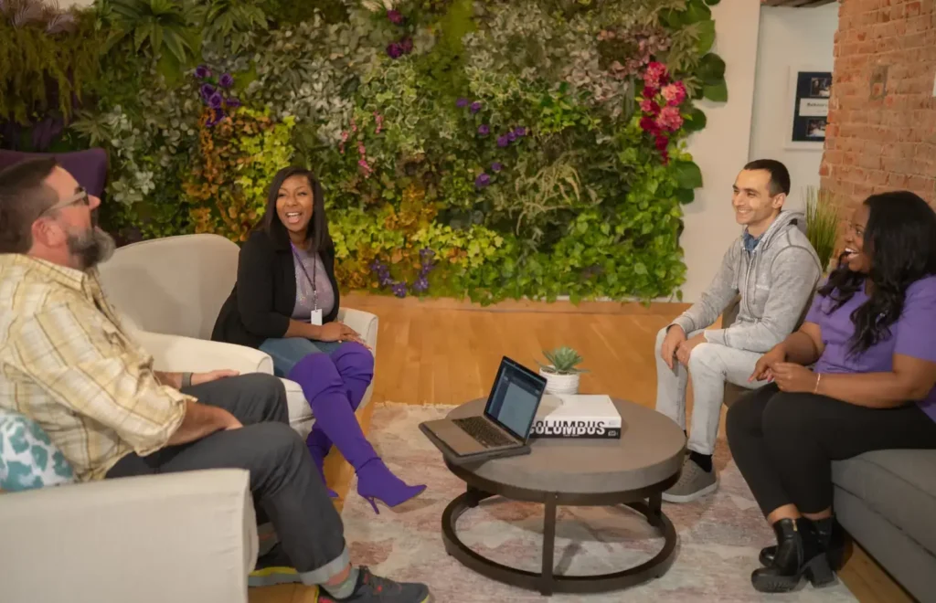 Coworkers having a casual group discussion in a modern office lounge with a green plant wall.