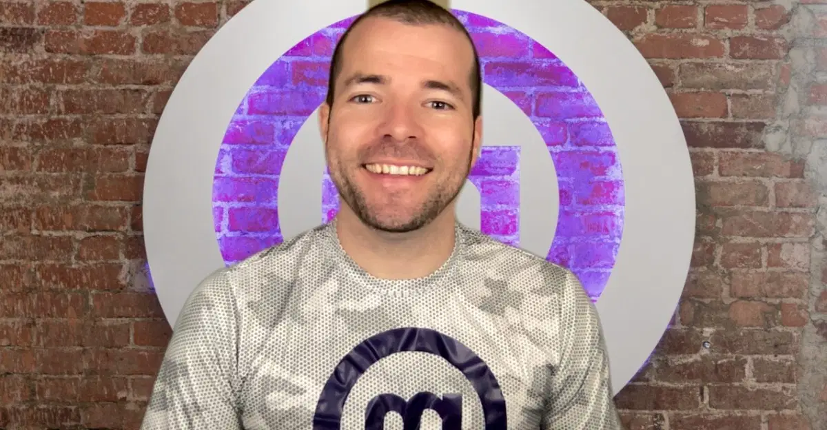Smiling man wearing a MentorcliQ shirt in front of a brick wall with the company logo.