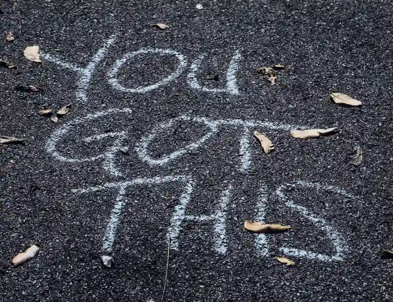 Close-up of chalk writing on asphalt saying 'YOU GOT THIS' surrounded by scattered leaves, conveying an encouraging message.